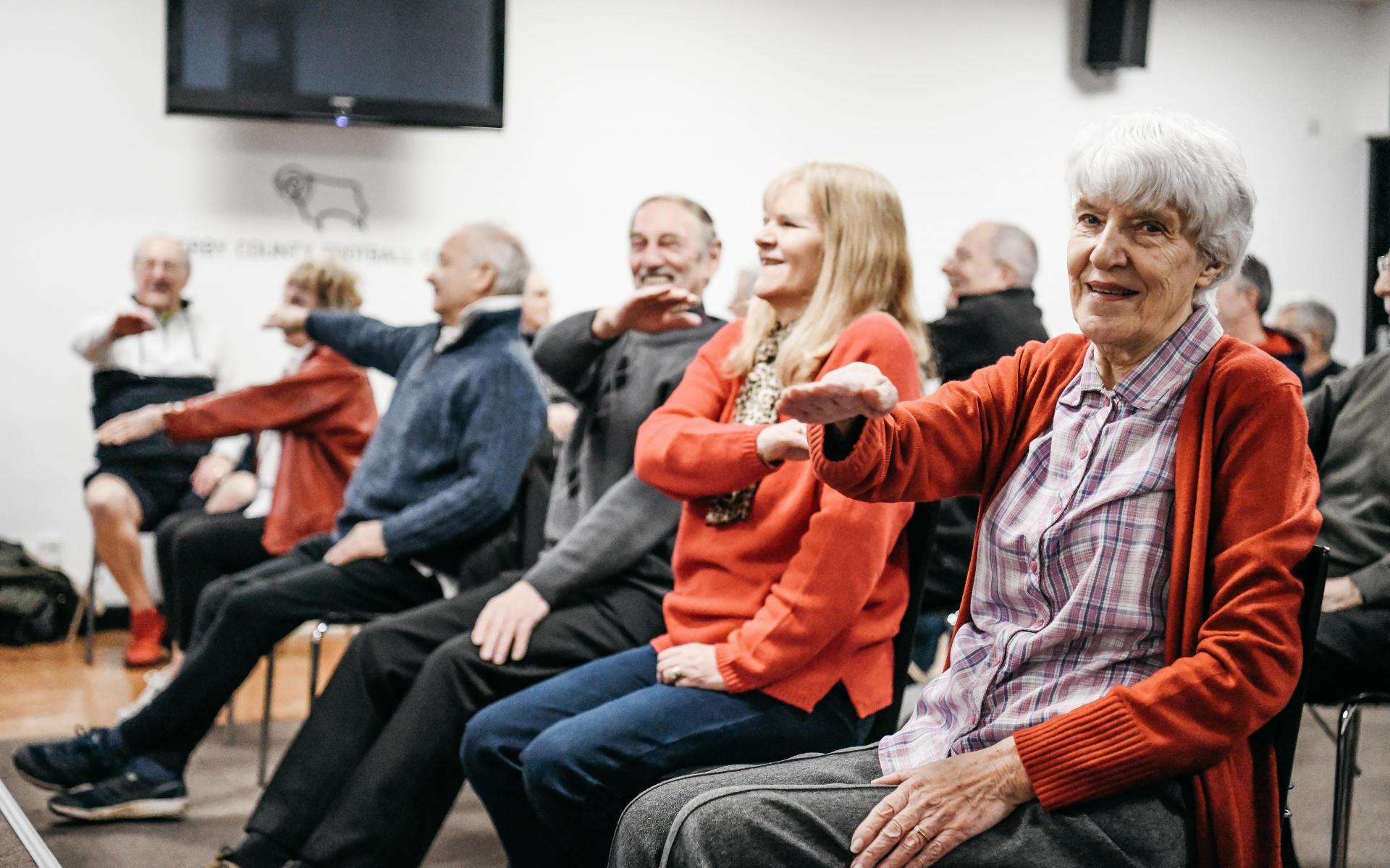 Elderly men and women sitting in chairs raising their hands doing exercise.