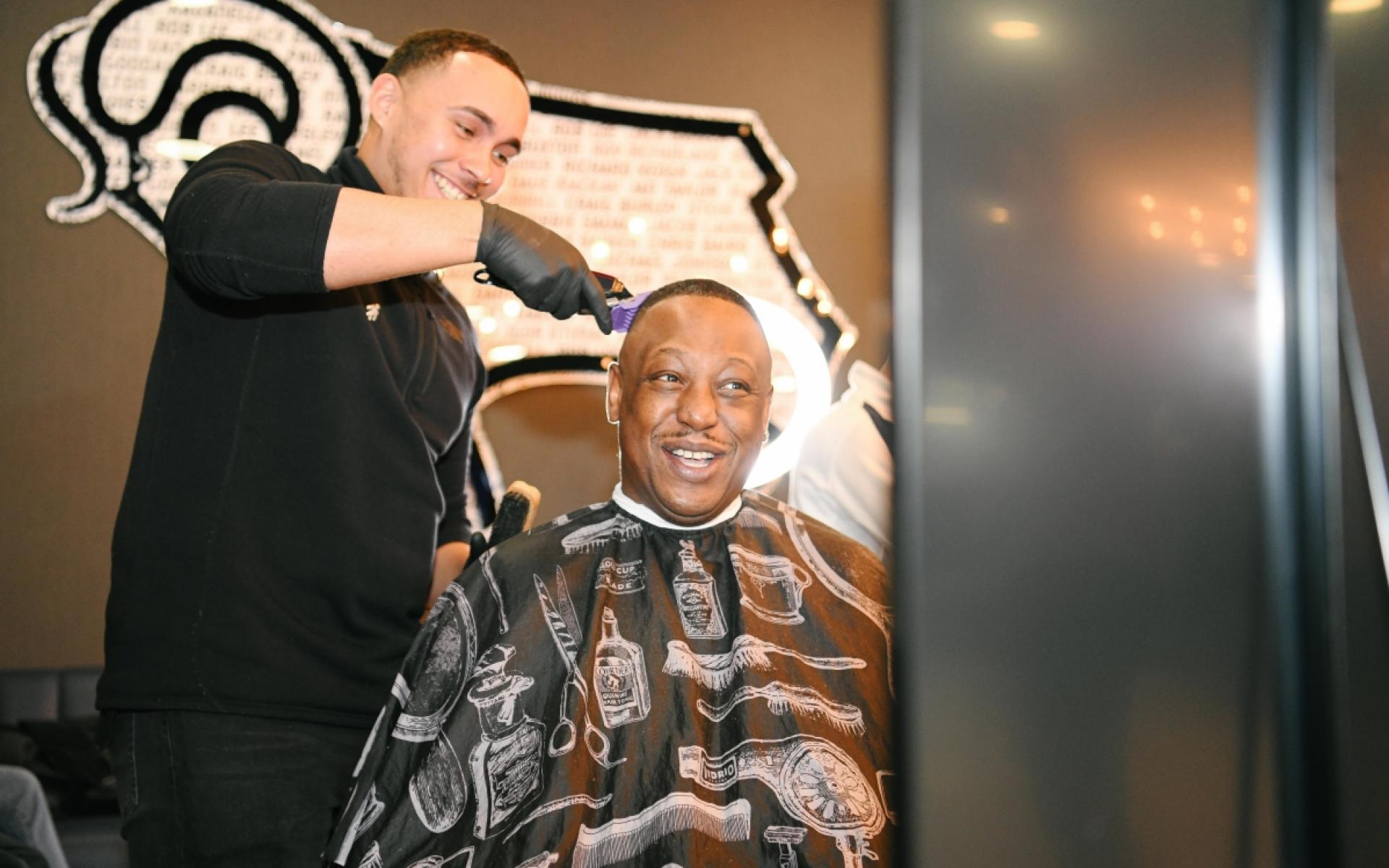 Barber shaving another man's head at pride park stadium, both smiling.