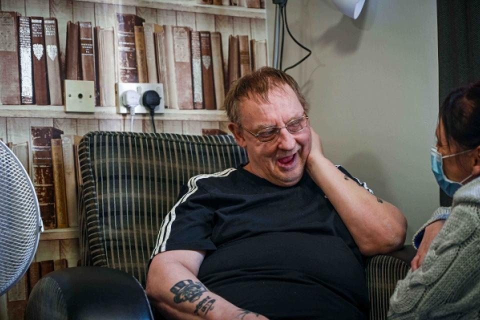Man sat in a chair in a care home lounge smiling at the camera